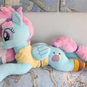 May include: A large plush toy pony in a lying position. The pony is primarily light blue with pink hair and tail. It has a yellow chest and a design of yarn and needles on its side. The toy is on a patterned surface.