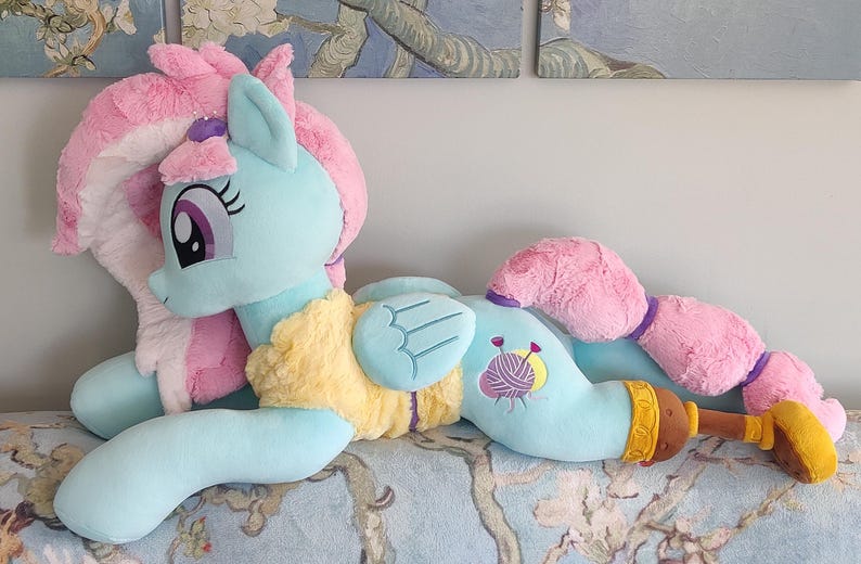 May include: A large plush toy pony in a lying position. The pony is primarily light blue with pink hair and tail. It has a yellow chest and a design of yarn and needles on its side. The toy is on a patterned surface.
