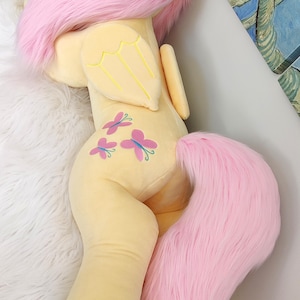 Puede incluir: Un gran juguete de peluche de un pony amarillo con pelo y cola rosas. El pony tiene una corona de flores en la cabeza y alas de mariposa en la espalda. El pony est&aacute; acostado de lado con la cabeza apoyada en su pata delantera.