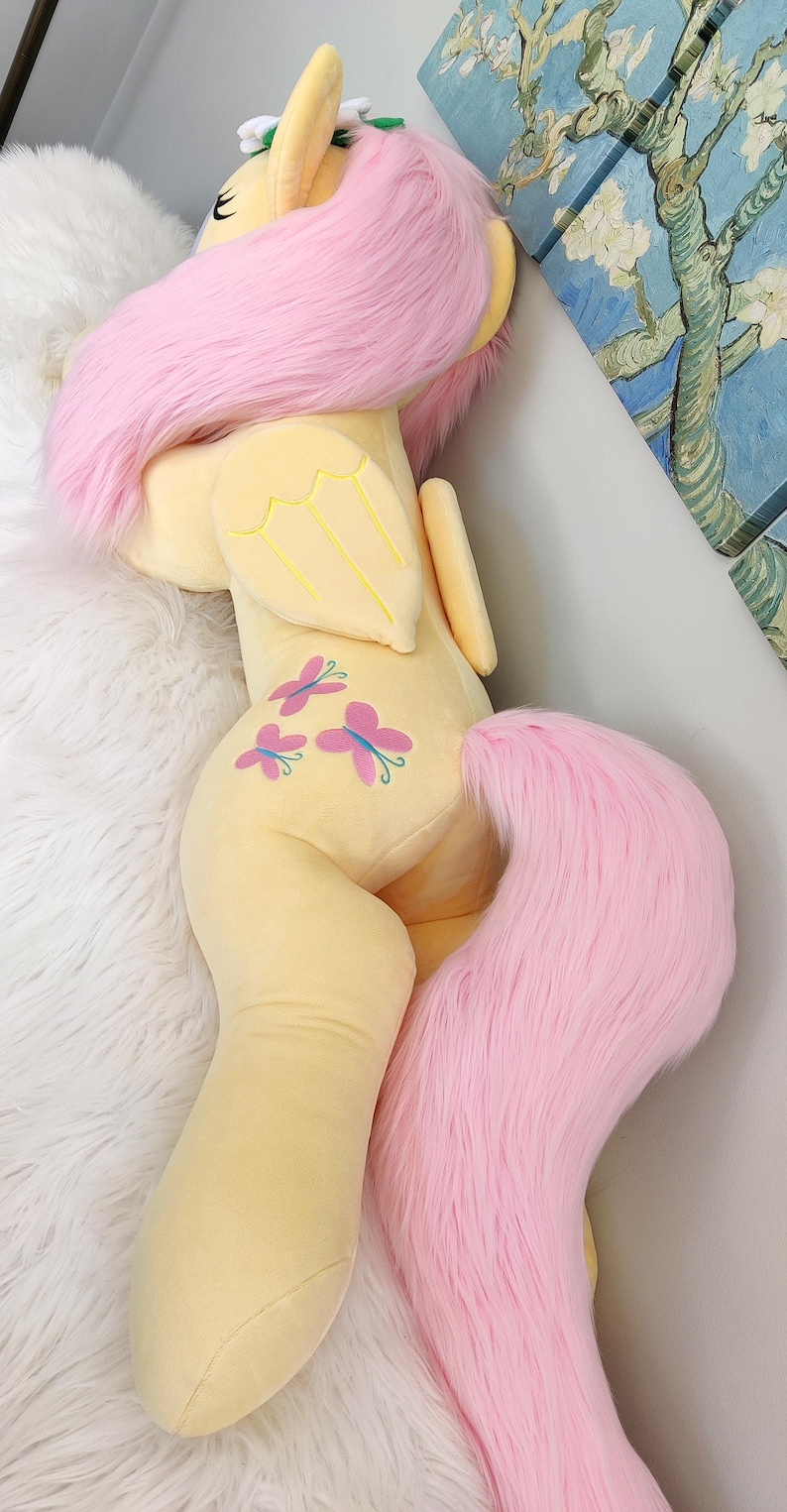 Puede incluir: Un gran juguete de peluche de un pony amarillo con pelo y cola rosas. El pony tiene una corona de flores en la cabeza y alas de mariposa en la espalda. El pony est&aacute; acostado de lado con la cabeza apoyada en su pata delantera.