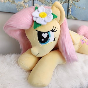 Puede incluir: Un gran juguete de peluche de Pinkie Pie de My Little Pony, acostado con la cabeza apoyada en sus patas delanteras. Tiene una melena y una cola rosadas, un cuerpo amarillo y una margarita blanca con un centro amarillo en la cabeza.