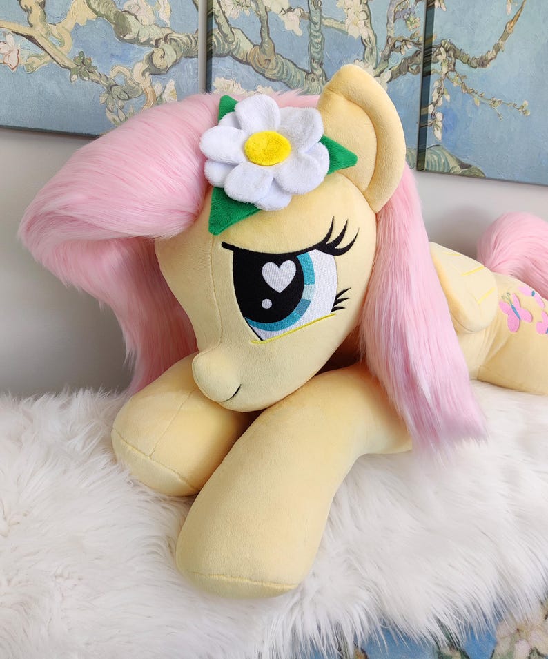 Puede incluir: Un gran juguete de peluche de Pinkie Pie de My Little Pony, acostado con la cabeza apoyada en sus patas delanteras. Tiene una melena y una cola rosadas, un cuerpo amarillo y una margarita blanca con un centro amarillo en la cabeza.
