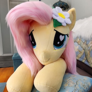 Puede incluir: Un gran juguete de peluche de Pinkie Pie de My Little Pony, con una melena y cola rosadas, un cuerpo amarillo y una flor blanca en la cabeza. El juguete est&aacute; tumbado sobre una almohada con estampado azul y blanco.