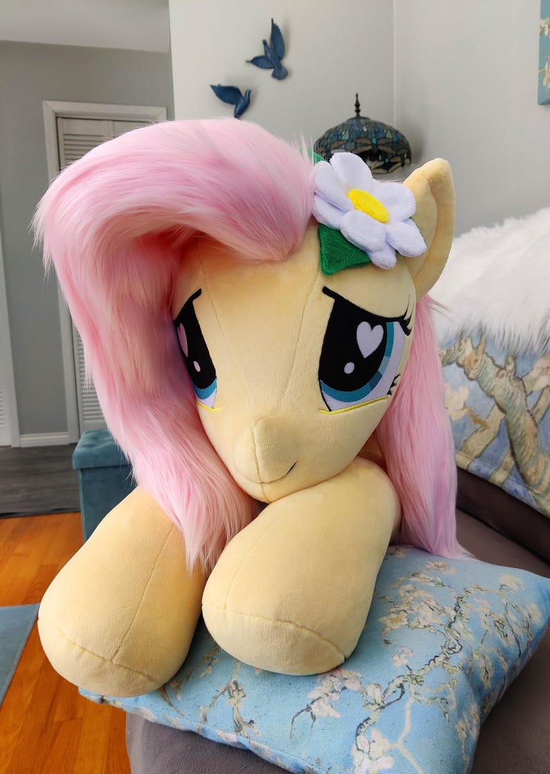 Puede incluir: Un gran juguete de peluche de Pinkie Pie de My Little Pony, con una melena y cola rosadas, un cuerpo amarillo y una flor blanca en la cabeza. El juguete est&aacute; tumbado sobre una almohada con estampado azul y blanco.