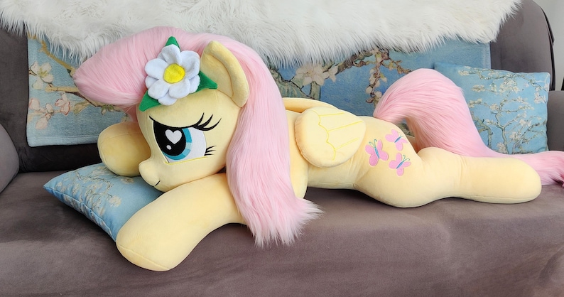 Puede incluir: Un juguete de peluche de Fluttershy de My Little Pony, acostado sobre una almohada. Es de color amarillo claro con una melena y cola rosadas. Tiene una flor en su melena y dise&ntilde;os de mariposas en su flanco.