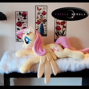 Puede incluir: Un juguete de peluche de Pinkie Pie de My Little Pony, acostado sobre una superficie blanca y esponjosa. El juguete es amarillo con pelo y alas rosas, y tiene una corona de flores en la cabeza. El juguete tiene un dise&ntilde;o de mariposa rosa en un lado.