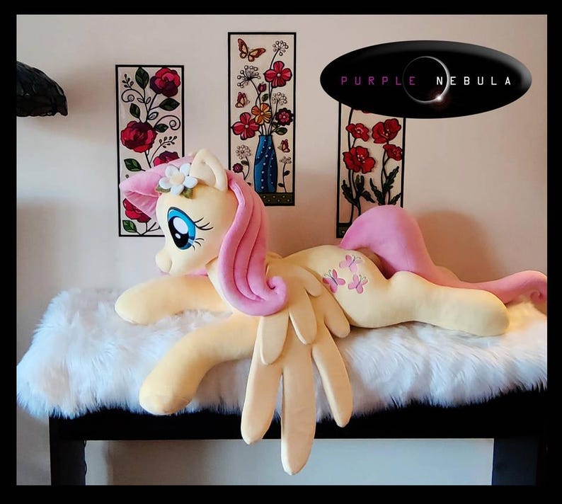 Puede incluir: Un juguete de peluche de Pinkie Pie de My Little Pony, acostado sobre una superficie blanca y esponjosa. El juguete es amarillo con pelo y alas rosas, y tiene una corona de flores en la cabeza. El juguete tiene un dise&ntilde;o de mariposa rosa en un lado.