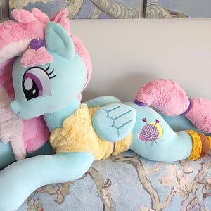 May include: A light blue plush pony toy with pink and yellow details. The pony features a long, pink mane and tail, and large eyes. It is lying on a patterned surface.
