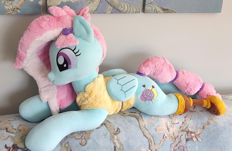 May include: A light blue plush pony toy with pink and yellow details. The pony features a long, pink mane and tail, and large eyes. It is lying on a patterned surface.