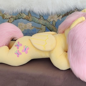 Puede incluir: Un gran juguete de peluche de un pony rosa con cuerpo amarillo y alas. El pony tiene una melena y una cola rosas y est&aacute; tumbado de lado con la cabeza apoyada en las patas delanteras. El pony tiene tres dise&ntilde;os de mariposas rosas en el costado.