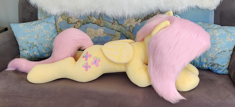 Puede incluir: Un gran juguete de peluche de un pony rosa con cuerpo amarillo y alas. El pony tiene una melena y una cola rosas y est&aacute; tumbado de lado con la cabeza apoyada en las patas delanteras. El pony tiene tres dise&ntilde;os de mariposas rosas en el costado.
