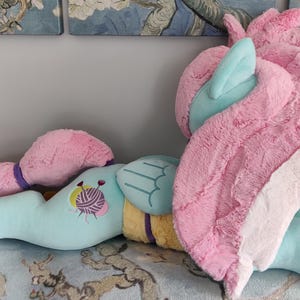 May include: A light blue unicorn-shaped cushion with pink and white wings, mane, and tail. The cushion has a yellow and purple accent and a yarn ball design. The toy is lying on a patterned surface.