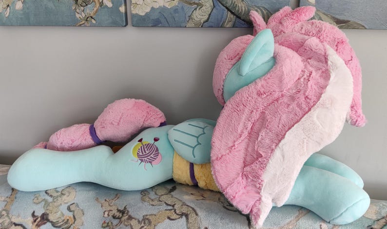 May include: A light blue unicorn-shaped cushion with pink and white wings, mane, and tail. The cushion has a yellow and purple accent and a yarn ball design. The toy is lying on a patterned surface.