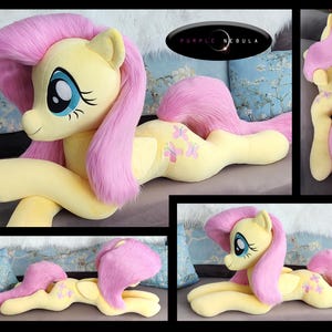 Puede incluir: Un gran juguete de peluche de Fluttershy de My Little Pony. El juguete es amarillo con pelo rosa y una cola rosa. Est&aacute; tumbado en un sof&aacute; con la cabeza apoyada en las patas delanteras. El juguete tiene un dise&ntilde;o de mariposa rosa en un lado.