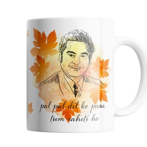 Könnte beinhalten: Weiße Keramiktasse mit einer Schwarzweißillustration eines Mannes vor einem Aquarellhintergrund aus orangefarbenen und gelben Blättern. Der Text "pal pal dil ke paas, tum raheti ho" ist auf die Tasse gedruckt.