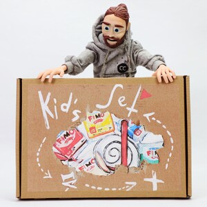Könnte beinhalten: Ein brauner Karton mit der Aufschrift "Kid's Set" und einer Zeichnung verschiedener Bastelartikel. Eine Puppenfigur in einem grauen Kapuzenpullover hält die Box. Die Box hat einen eingerissenen Bereich.