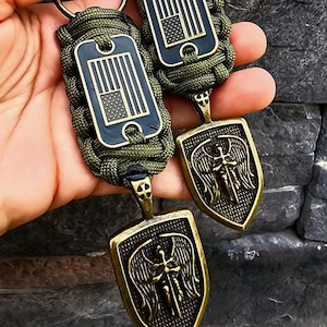 May include: Two keychains with green paracord and metal charms. One charm is a shield with an angel and sword design. The other charm is a rectangular tag with an American flag design.