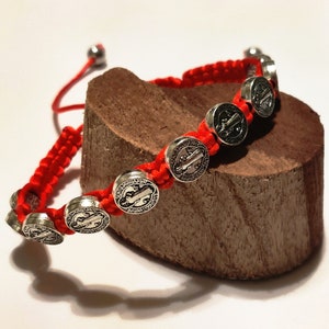 May include: Red string bracelet with silver medallions featuring Saint Benedict's image and prayer.