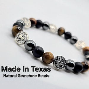 May include: A beaded bracelet featuring a mix of natural gemstone beads in brown, black, and silver tones. The bracelet includes decorative silver Saint Benedict medallions. The text "Made In Texas" and "Natural Gemstone Beads" is displayed.