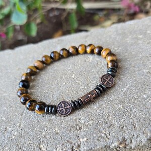 Puede incluir: Una pulsera de cuentas con piedras de ojo de tigre y cuentas negras. La pulsera incluye una cuenta cil&iacute;ndrica de color cobre con una cruz y dos cuentas redondas con un emblema religioso.