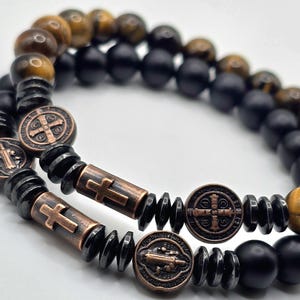 Peut inclure: Deux bracelets de perles. L'un pr&eacute;sente des perles &oelig;il de tigre et noires, l'autre des perles noires. Les deux bracelets ont des accents de croix et de m&eacute;daillon de couleur bronze. Les bracelets sont sur une surface blanche.