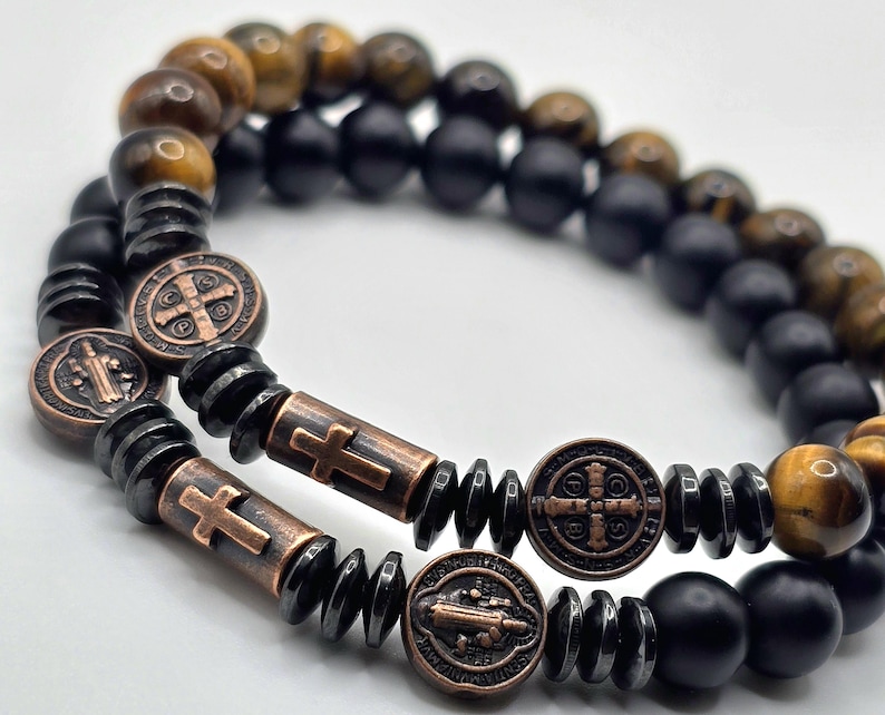 Peut inclure: Deux bracelets de perles. L'un pr&eacute;sente des perles &oelig;il de tigre et noires, l'autre des perles noires. Les deux bracelets ont des accents de croix et de m&eacute;daillon de couleur bronze. Les bracelets sont sur une surface blanche.