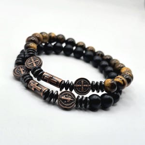 Peut inclure: Deux bracelets de perles superpos&eacute;s. L'un est compos&eacute; de perles &oelig;il de tigre et noires. L'autre pr&eacute;sente des perles noires, des croix et des m&eacute;daillons en bronze.
