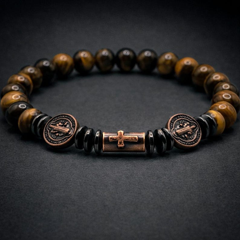 Puede incluir: Una pulsera de cuentas con piedras de ojo de tigre y detalles en metal oscuro. La pulsera incluye dos piezas decorativas redondas de color cobre y una pieza rectangular con un dise&ntilde;o de cruz. Las cuentas tienen aproximadamente 0,8 cm de di&aacute;metro.