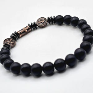 Peut inclure: Bracelet de perles noires avec des perles d'onyx mat et des accents m&eacute;talliques couleur bronze. Le bracelet comprend une croix et deux m&eacute;daillons ronds avec des symboles religieux. Les perles mesurent environ 8 mm de diam&egrave;tre.
