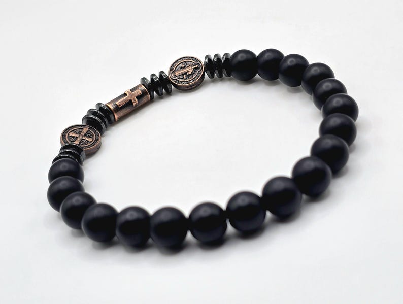 Peut inclure: Bracelet de perles noires avec des perles d'onyx mat et des accents m&eacute;talliques couleur bronze. Le bracelet comprend une croix et deux m&eacute;daillons ronds avec des symboles religieux. Les perles mesurent environ 8 mm de diam&egrave;tre.
