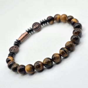 Puede incluir: Pulsera de cuentas de ojo de tigre, con elementos marrones, negros y cobrizos. El dise&ntilde;o incluye un cierre y elementos decorativos. La pulsera se muestra sobre un fondo blanco. Las cuentas tienen aproximadamente 0,8 cm de di&aacute;metro.