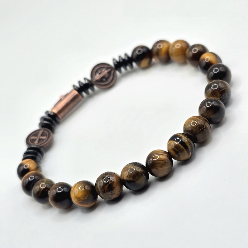 Puede incluir: Pulsera de cuentas de ojo de tigre, con elementos marrones, negros y cobrizos. El dise&ntilde;o incluye un cierre y elementos decorativos. La pulsera se muestra sobre un fondo blanco. Las cuentas tienen aproximadamente 0,8 cm de di&aacute;metro.