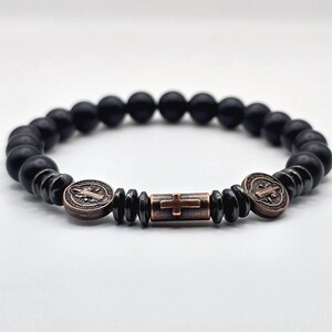 Peut inclure: Bracelet de perles noires avec des accents de couleur bronze. Le bracelet pr&eacute;sente un motif de croix central et deux m&eacute;daillons circulaires avec des d&eacute;tails en relief. Les perles sont mates et les &eacute;l&eacute;ments m&eacute;talliques ont une finition vintage.
