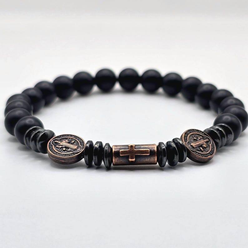 Peut inclure: Bracelet de perles noires avec des accents de couleur bronze. Le bracelet pr&eacute;sente un motif de croix central et deux m&eacute;daillons circulaires avec des d&eacute;tails en relief. Les perles sont mates et les &eacute;l&eacute;ments m&eacute;talliques ont une finition vintage.