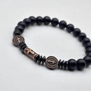 Peut inclure: Bracelet de perles noires avec des &eacute;l&eacute;ments crois&eacute;s et m&eacute;daillons de couleur bronze. Le bracelet est compos&eacute; de perles noires mates et a un design extensible. Les accents en bronze comprennent une croix et des m&eacute;daillons circulaires grav&eacute;s.