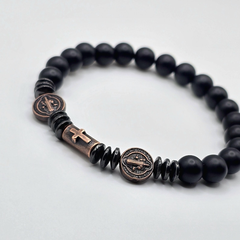 Peut inclure: Bracelet de perles noires avec des &eacute;l&eacute;ments crois&eacute;s et m&eacute;daillons de couleur bronze. Le bracelet est compos&eacute; de perles noires mates et a un design extensible. Les accents en bronze comprennent une croix et des m&eacute;daillons circulaires grav&eacute;s.
