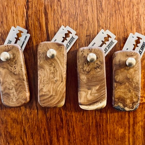 May include: Five wooden holders for razor blades. Each holder is made of a dark brown wood with a unique grain pattern. The holders have a small metal knob on the top and a silver razor blade attached to the front. The razor blades are labeled "Derby Extra".