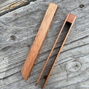 Puede incluir: Dos pinzas de cocina de madera, una está cerrada y la otra está abierta. Las pinzas son de madera marrón claro con un tono rojizo.