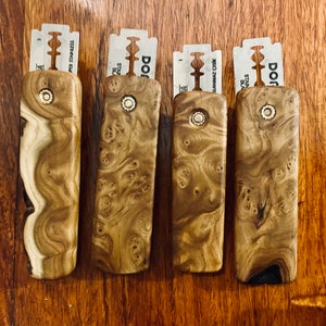 May include: Four wooden razor holders with stainless steel blades. The holders have unique wood grain patterns and a brass-colored accent. The blades are labeled with the brand name "DORCO". The items are displayed on a wooden surface.