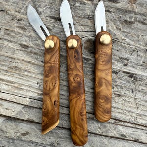 May include: Three small, folding pocket knives with wooden handles. The handles are made of a dark brown wood with a unique grain pattern. The blades are silver and have a slight curve.