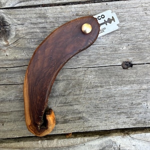 May include: A handcrafted, curved wooden razor handle with a brass rivet. A silver razor blade is inserted into the handle. The wood grain is visible, showing a mix of dark and light brown tones. The blade has the text "CO" and "STAINLESS BLADE".