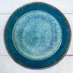 May include: A handcrafted ceramic plate with a turquoise glaze. The plate features a raised, textured rim with a rope-like design. The center of the plate has a lighter blue hue, creating a layered effect. This decorative plate is suitable for serving food.