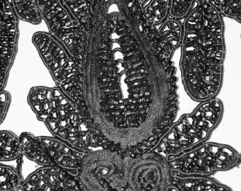 Può includere: Primo piano di un tessuto nero con perline con intricati motivi floreali e astratti. Il design presenta una forma centrale a goccia e dettagli a spirale, il tutto su sfondo bianco. Le perline creano un effetto strutturato e dimensionale.