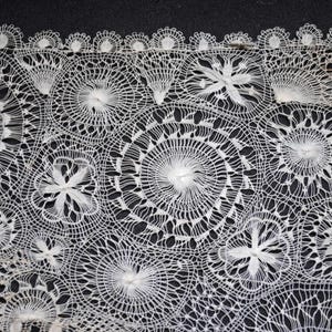 May include: Close-up of white lace fabric with intricate floral and circular patterns. The lace features detailed openwork designs, creating a delicate and textured appearance. The lace is set against a dark background, highlighting the detailed patterns.