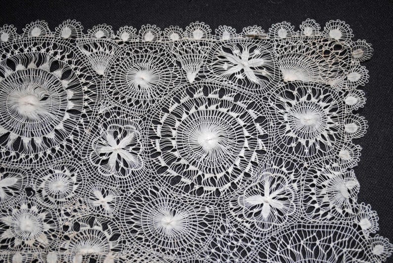 May include: Close-up of white lace fabric with intricate floral and circular patterns. The lace features detailed openwork designs, creating a delicate and textured appearance. The lace is set against a dark background, highlighting the detailed patterns.