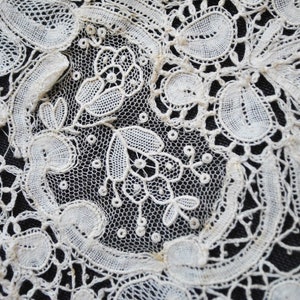 May include: Close-up of intricate white lace with floral and leaf patterns. The delicate design features openwork and detailed stitching, creating a textured surface. The lace is set against a dark background, highlighting the fine details.