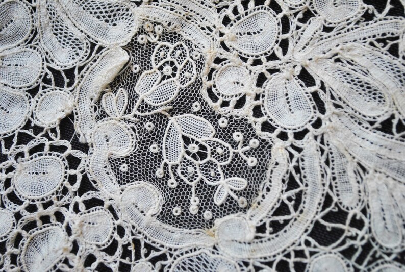 May include: Close-up of intricate white lace with floral and leaf patterns. The delicate design features openwork and detailed stitching, creating a textured surface. The lace is set against a dark background, highlighting the fine details.