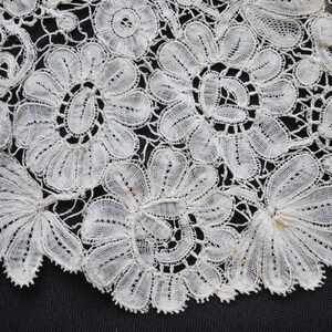 May include: Close-up of delicate white lace with floral and leaf patterns. The lace features intricate details and a scalloped edge, creating a textured and elegant design. The lace is likely used for decorative purposes in clothing or home decor.