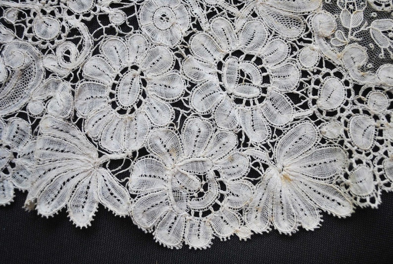 May include: Close-up of delicate white lace with floral and leaf patterns. The lace features intricate details and a scalloped edge, creating a textured and elegant design. The lace is likely used for decorative purposes in clothing or home decor.