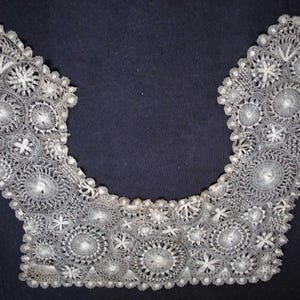 May include: A delicate, white lace collar with an intricate floral and circular pattern. The collar is U-shaped, with detailed openwork and scalloped edges. The lace is set against a dark background.